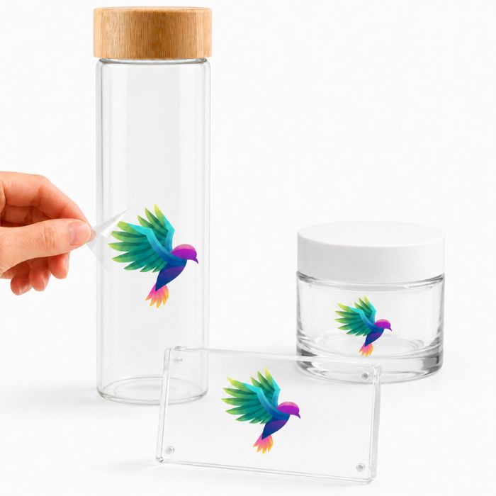 Pegatina transparente personalizada despegándose de una botella de cristal junto a tarro cosmético y metacrilato